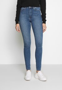 Jeans ajustados de denim en color azul con una cintura alta. Presenta cinco pasadores para el cinturón y costuras clásicas. Combinados con zapatillas blancas.