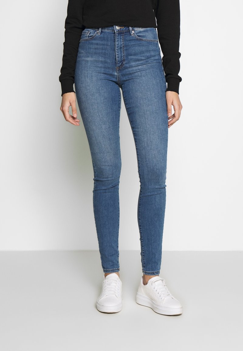 Jeans ajustados de denim en color azul con una cintura alta. Presenta cinco pasadores para el cinturón y costuras clásicas. Combinados con zapatillas blancas.