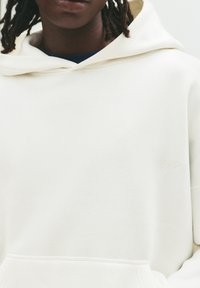 Weißer Hoodie aus weichem Material, mit einer Kängurutasche, verstellbarem Kapuzenpullover und einem dezenten gestickten Logo auf der Brust.