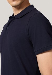 Granatowa koszulka polo z krótkim rękawem, z prążkowanym kołnierzykiem i zapięciem na dwa guziki, wykonana z fakturowanej tkaniny. Zbliżenie na ramię.