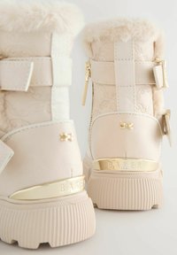 Stivali beige chiaro con risvolti in pelliccia, fibbie e un accento dorato con la scritta "BAKER" impressa sul lato.
