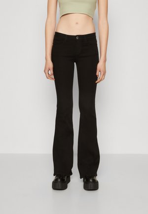 Kvinde iført sorte udvidede jeans, sorte platformssko og en cropped lysegrøn top, stående mod en ensfarvet hvid baggrund.