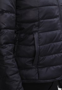 Sort puffjakke med en quiltet design, der har en sidelomme og en glat, shiny stofstruktur. Accenter inkluderer en skjult lynlås.