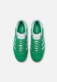 Sapatilhas verdes em camurça com detalhes brancos, apresentando três riscas, atacadores redondos e uma sola de borracha texturizada. Logótipo da Adidas na língua.