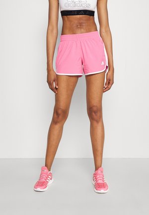 Personne portant un short de sport rose Adidas, un crop top blanc avec le logo Adidas et des chaussures de sport roses sur un fond uni.