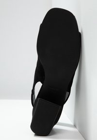Semelle d’une chaussure à talon noir, face à la caméra, appuyée en angle contre un coin de mur et de sol blancs.
