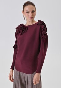 Maglione a maglia color bordeaux con dettagli arruffati sulle maniche, scollatura rotonda e vestibilità rilassata, abbinato a una gonna plissettata a quadri.