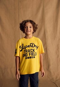 Geel katoenen t-shirt met zwart opdruk "SuperDry TRACK AND FIELD", korte mouwen en een relaxte pasvorm. Zwarte cargoshorts worden eronder gedragen.