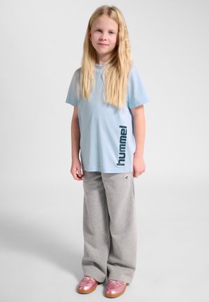 Blondynka stojąca, ubrana w jasnoniebieską koszulkę Hummel, szare szerokie spodnie i błyszczące różowe buty na neutralnym tle.