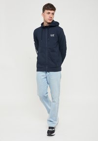 Felpa con zip di colore navy, dotata di tasca frontale, cappuccio con coulisse e logo bianco sul petto; abbinata a jeans azzurri chiari e sneakers nere.