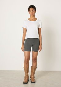 Bela kratka majica s kratkimi rokavi, sivo vzorčaste kratke hlače in rjavi usnjeni škornji do kolen s sponkami. Model stoji na nevtralnem ozadju tal.