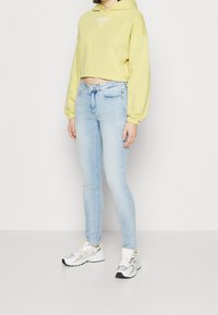 Un hoodie recortado de color amarillo claro con un logo, combinado con jeans ajustados de color azul claro y zapatillas blancas con acentos amarillos y detalles grises.