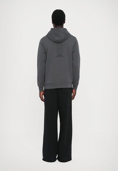 Grijze hoodie met een capuchon en een voorzak, met geprinte tekst op de achterkant. Gecombineerd met losse zwarte broek. Zachte stof textuur.