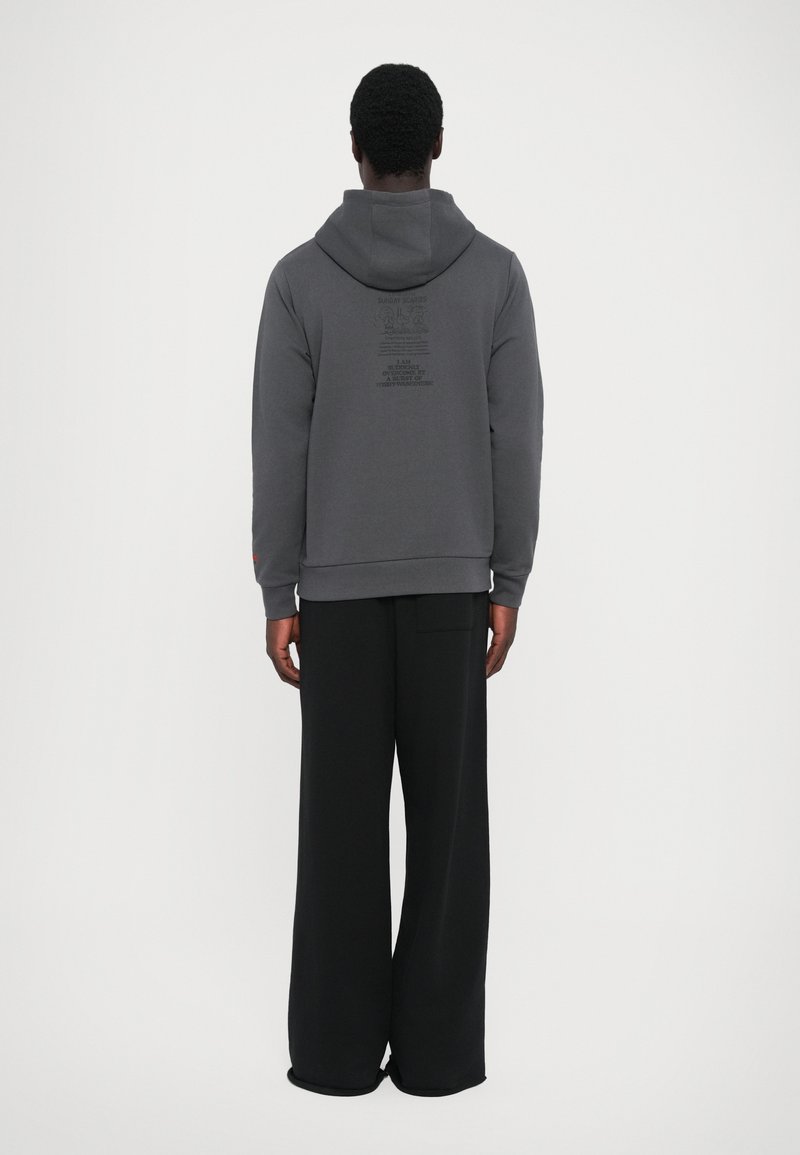 Grau Hoodie mit einer Kapuze und einer Fronttasche, auf der Rückseite mit bedrucktem Text. Kombiniert mit lockeren schwarzen Hosen. Weicher Stoff.