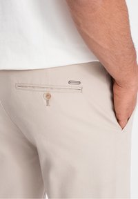 Beżowe spodnie z tylnią kieszenią zapinaną na guzik i biała koszula, ukazująca prawą dłoń spoczywającą w przedniej kieszeni.