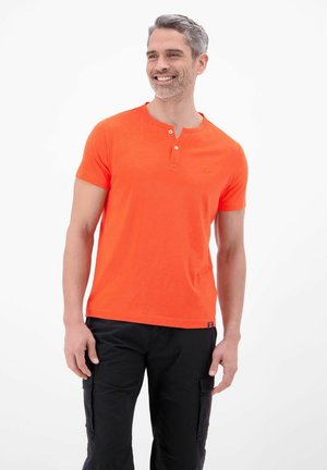 Lächelnder Mann mit kurzem grauen Haar, der ein orangenes kurzärmeliges Henley-Shirt und schwarze Cargohosen trägt, steht vor weißem Hintergrund.