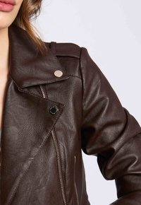 Veste en cuir synthétique marron de style motard avec une fermeture à glissière asymétrique, des finitions en argent, des revers larges et une surface texturée.