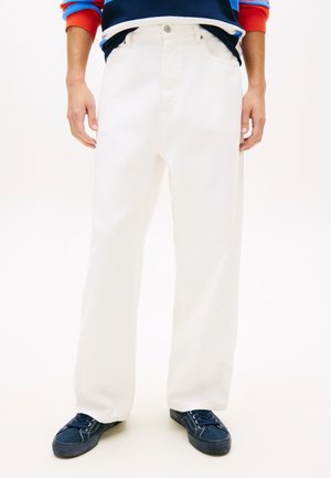 Mand iført løse hvide bukser, marineblå sneakers og en flerfarvet sweater med orange manchetter, stående på en ensfarvet hvid baggrund.