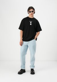 Schwarzes, übergroßes T-Shirt mit weißem "REPLAY"-Logo und Grafik; hellblaue Jeans; schwarze klobige Sneaker; Model trägt dunkle Sonnenbrillen.
