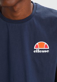 Tengeri kék pamut kereknyakú póló, fehér "ellesse" logóval és színes narancssárga-fehér félkör alakú grafikával a mellrészen.