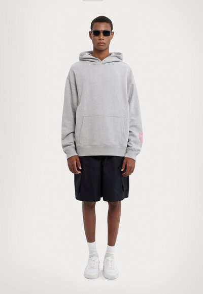 Sweatshirt gris à capuche avec poche avant, shorts noirs, baskets blanches et lunettes de soleil noires. Présente un graphique rose sur la manche.