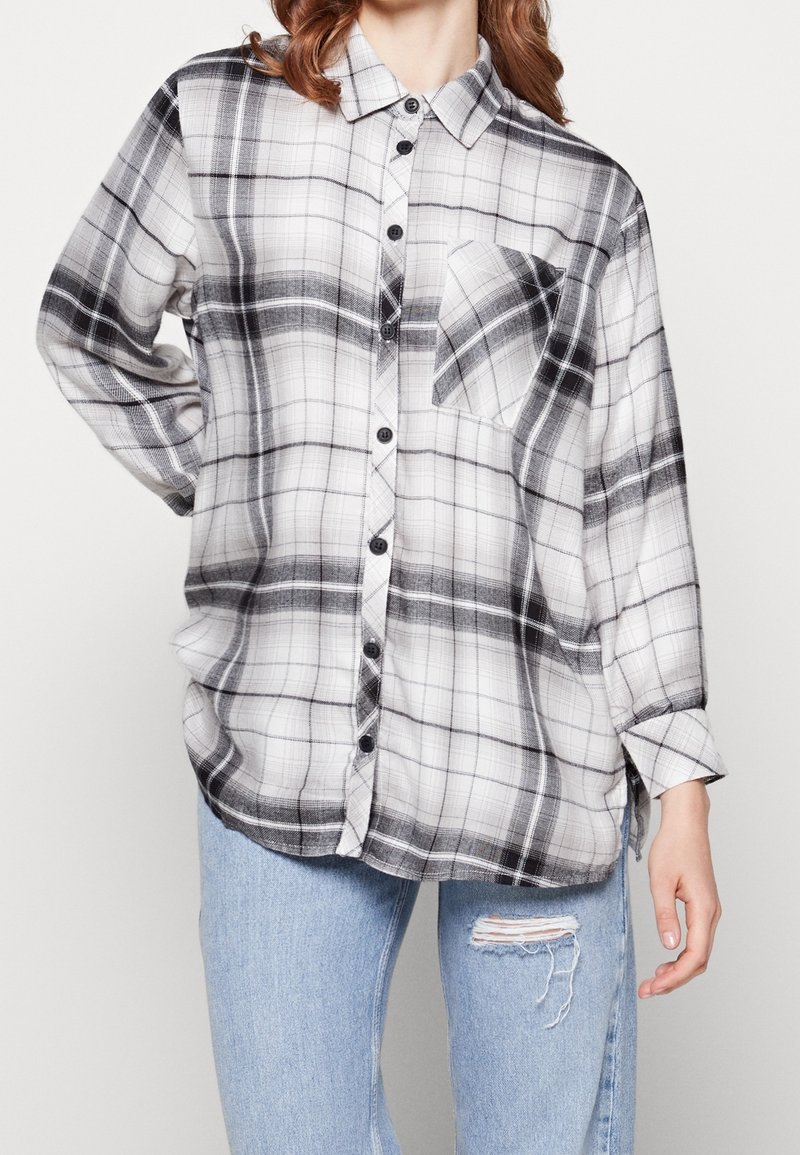 Femme portant une chemise ample à carreaux noir et blanc avec une poche poitrine, associée à un jean bleu clair déchiré.