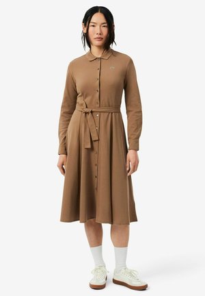 Mujer con vestido marrón de manga larga con botones y cinturón en la cintura, calcetines blancos y zapatillas color crema, de pie frente a un fondo liso.