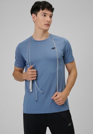 Hombre con camiseta deportiva azul y pantalones negros sosteniendo una cuerda para saltar colgada alrededor de su cuello, mirando hacia un lado.