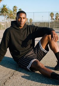 Jonge man in zwarte hoodie en gestreepte korte broek zittend op een buitenbasketbalveld met palmbomen en een hek op de achtergrond.