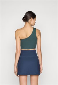 Roheline ühes õlaga lühike topp, millel on vormiv disain, kombineeritud tumesinise seelikuga. Sile kangas, millel on õmbluse detail taljes.