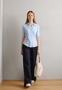 Chemise boutonnière bleu clair à manches courtes associée à un pantalon large en denim foncé. Tenant un sac fourre-tout couleur crème ; fond neutre.