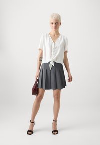 Weiße Kurzarmbluse mit Knotenfront, kombiniert mit einem grauen, plissierten Rock. Das Modell trägt eine bordeauxfarbene Tasche und schwarze Sandalen mit Absatz.