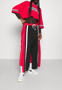 Rotes, kurzes Sportshirt mit bedrucktem Logo, kombiniert mit schwarzen weit geschnittenen Hosen mit roten und weißen Seitenstreifen und verstellbarem Kordelzug.