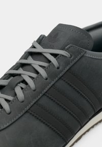 Γκρι δερμάτινο αθλητικό παπούτσι με τρεις πιο σκούρες γκρι ρίγες, γκρι κορδόνια και υφασμένο λαστιχένιο πέλμα. Το λογότυπο στη γλώσσα του παπουτσιού είναι ορατό.