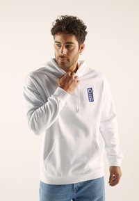 White hoodie with a front pocket and drawstring hood. Features a blue patch with stylized text. Pairing with light blue jeans.