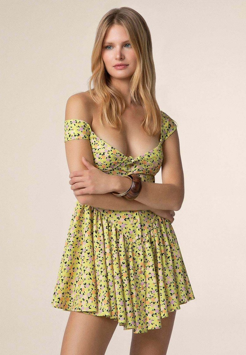 Abito floreale giallo con maniche scoperte, gonna a pieghe e design con nodo frontale. Presenta un motivo astratto colorato.