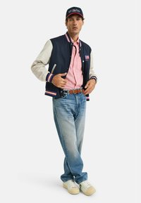 Giacca varsity blu navy con maniche color crema, camicia a righe rossa e bianca, jeans azzurri chiari e sneaker bianche con accenti color crema. Cappello scuro.