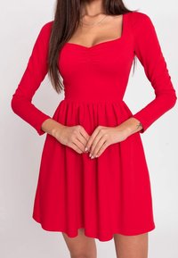 Robe rouge avec des manches longues et un décolleté carré. Le buste présente un plissage horizontal, avec une jupe évasée tombant juste au-dessus des genoux.