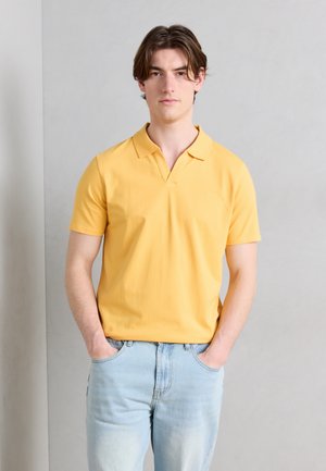 Młody mężczyzna z rękami w kieszeniach, ubrany w żółtą koszulkę polo z krótkim rękawem i jasnoniebieskie dżinsy, na jednolitym jasnoszarym tle.