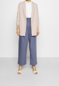 Blazer beige oversize, chemise blanche boutonnée et pantalon large bleu marine à texture lisse. Associé à des sandales à talons blanc cassé.