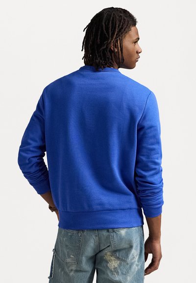 Zils džemperis ar apaļu kakla izgriezumu, ribotām piedurknēm un apakšmalu. Izgatavots no mīksta auduma ar gludu tekstūru. Nēsā virs nolietotām džinsu biksēm.