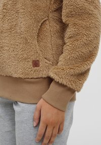 Tan fliisist pullover, millel on pehme tekstuur, ribilised pruunid mansetid ja allääre osa, ning väike pruun brändi silt. Hallid dressipüksid on osaliselt nähtavad.