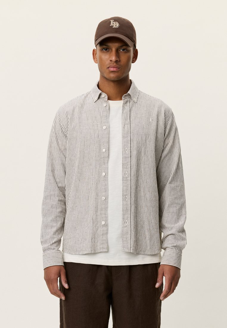 Ριγέ πουκάμισο με κουμπιά σε ανοιχτό γκρι και λευκό ύφασμα, φορεμένο πάνω από ένα λευκό T-shirt. Συμπληρώνεται με καφέ καπέλο και σκούρα καφέ παντελόνια.