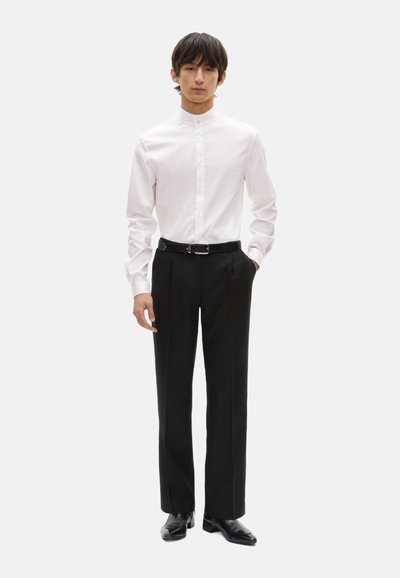 Weiß gestreifte Hemdbluse mit Stehkragen, kombiniert mit schwarzen plissierten Hosen und schwarzen Schuhen. Glattes Material, taillierte Passform.