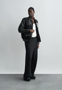 Fekete varsity dzseki bőr ujjal, fehér felirattal és bordázott mandzsettákkal. Széles fekete nadrággal és fehér pólóval párosítva.