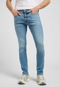 Mann iført slim-fit lyseblå jeans og hvite sneakers, stående med hendene i lommene mot en ensfarget grå bakgrunn.