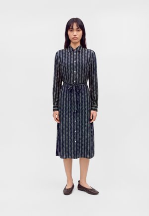 Femme portant une robe noire à manches longues boutonnée avec des motifs verticaux blancs en forme de chaîne et des chaussures plates foncées, debout devant un fond uni.