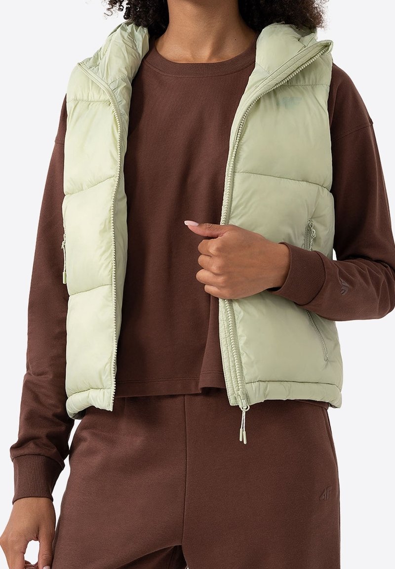 Personne portant une chemise marron à manches longues et un pantalon avec une veste matelassée sans manches vert clair, main tenant la fermeture éclair de la veste près de la poitrine.