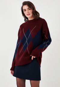 Bordo in mornarsko modro podrobljeno pulover iz mehkega pletenega materiala, ki ima sproščen kroj in visok ovratnik. Oblečen z temnimi krili.