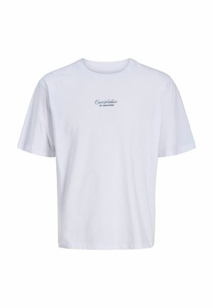 Młoda osoba stoi i się uśmiecha, ubrana w biały t-shirt z nadrukiem, czarne szorty w cienkie paski, białe skarpetki z logo i białe trampki.